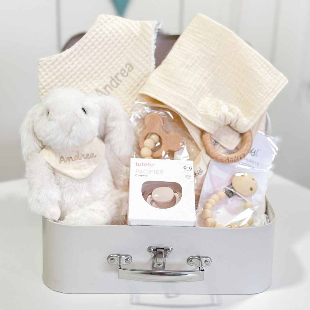 Canastilla bebé Bunny compuesta por peluche conejito con bandana personalizada con el nombre del bebé, chupetero de silicona modelo Momma personalizado, mordedor de silicona con figura de madera natural haya, chupete retro nordic personalizado, doudou muselina con aro de madera natural de haya personalizado, bandana de waffle algodón personalizado y neceser de waffle algodón personalizado. Todos los productos se presentan en una bonita y práctica maletita también personalizada con el nombre del bebé.