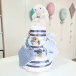 Tarta de pañales de tres pisos compuesto por 52 unidades marca Dodot, peluche principal conejito personalizado, sujeta chupetes de silicona, mordedor orejitas piqué waffle, algodón y madera personalizado, cepillo y peine para bebé de madera 100% natural, mordedor de silicona, bandana de piqué waffle y osito Dou Dou personalizado. Dimensiones altura 55cm x 26cm ancho. Color azul oscuro