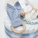 Tarta de pañales 1 piso LITTLE BUNNY compuesto por 28 unidades marca Dodot Sensitive T2, peluche principal conejito personalizado, mordedor de piqué waffle de algodón y aro de madera personalizado, sujeta chupetes de silicona modelo Momma personalizado y juego calcetines bebé de algodón. Dimensiones altura 35cm x 22cm ancho.