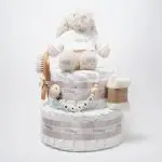 Tarta de pañales de dos pisos compuesta por 44 unidades marca Dodot Sensitive T2, peluche principal conejito personalizado, conjunto cepillo y peine bebé de madera personalizado, sujeta chupetes de silicona personalizado y juego calcetines bebé. Dimensiones altura 45cm x 26cm ancho. Color Beige/Toffe