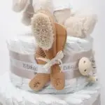 Tarta de pañales de dos pisos compuesta por 44 unidades marca Dodot Sensitive T2, peluche principal conejito personalizado, conjunto cepillo y peine bebé de madera personalizado, sujeta chupetes de silicona personalizado y juego calcetines bebé. Dimensiones altura 45cm x 26cm ancho. Color Beige/Toffe