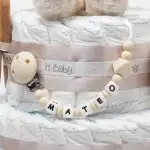 Tarta de pañales de dos pisos compuesta por 44 unidades marca Dodot Sensitive T2, peluche principal conejito personalizado, conjunto cepillo y peine bebé de madera personalizado, sujeta chupetes de silicona personalizado y juego calcetines bebé. Dimensiones altura 45cm x 26cm ancho. Color Beige/Toffe
