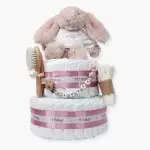 Tarta de pañales de dos pisos compuesta por 44 unidades marca Dodot Sensitive T2, peluche principal conejito personalizado, conjunto cepillo y peine bebé de madera personalizado con el nombre del bebé, sujeta chupetes de silicona personalizado con el nombre del bebé y juego calcetines bebé. Dimensiones altura 45cm x 26cm ancho. Color Rosa palo