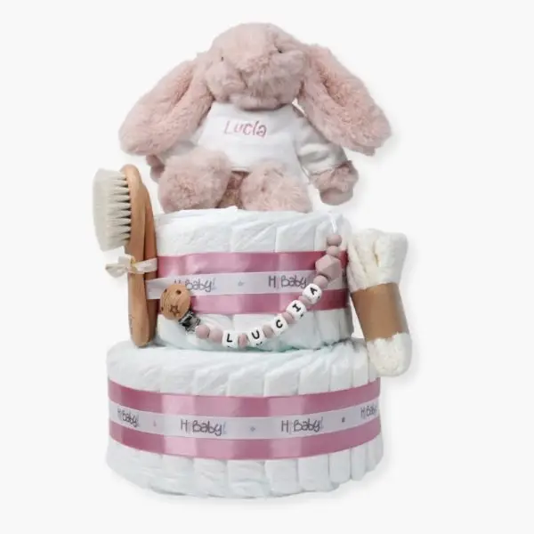 Tarta de pañales de dos pisos compuesta por 44 unidades marca Dodot Sensitive T2, peluche principal conejito personalizado, conjunto cepillo y peine bebé de madera personalizado con el nombre del bebé, sujeta chupetes de silicona personalizado con el nombre del bebé y juego calcetines bebé. Dimensiones altura 45cm x 26cm ancho. Color Rosa palo
