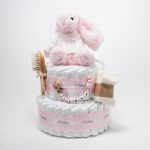 Tarta de pañales de dos pisos compuesta por 44 unidades marca Dodot Sensitive T2, peluche principal conejito personalizado, conjunto cepillo y peine bebé de madera personalizado con el nombre del bebé, sujeta chupetes de silicona personalizado con el nombre del bebé y juego calcetines bebé. Dimensiones altura 45cm x 26cm ancho. Color Rosa bebé