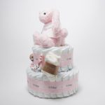 Tarta de pañales de dos pisos compuesta por 44 unidades marca Dodot Sensitive T2, peluche principal conejito personalizado, conjunto cepillo y peine bebé de madera personalizado con el nombre del bebé, sujeta chupetes de silicona personalizado con el nombre del bebé y juego calcetines bebé. Dimensiones altura 45cm x 26cm ancho. Color Rosa bebé
