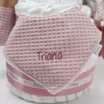 Tarta de pañales de 3 pisos compuesto por 52 unidades marca Dodot sensitive, pelota sensorial Montessori de Little Dutch, mordedor orejitas de tela piqué waffle algodón y aro de madera natural personalizado con el nombre del bebé, bandana de tela piqué waffle algodón personalizable, chupetero de silicona y crochet con pinza de madera natural de haya, paño de lactancia de piqué waffle algodón, muñeca ANNA de Little Dutch 35cm. Stick de madera personalizado con el nombre del bebé. Dimensiones altura 42cm x 27cm ancho. Color Rosa palo