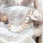 Tarta pañales V. Sunny Champagne compuesto por 84 unidades marca Dodot, peluche principal Mocca de Teddykompaniet 25 cm, chupetero de silicona de grado alimentario libre de BPA, PVC, Ftalatos, cadmio y plomo, bandana secababitas, chupete personalizado, mordedor orejitas y capa de baño de piqué waffle personalizada de Hi Baby, Doudou baby bunny personalizado, conjunto de cepillo y peine de madera personalizado hecho con materiales 100% naturales de lana de cabra y madera de haya, ofreciendo una experiencia cómoda y suave para tu bebé. Ideal para el cuero cabelludo sensible de los más pequeños, guardachupetes de silicona perfecto para guardar el chupete cuando no se está usando, body de babidu personalizado con el dibujo de un conejito y el nombre del bebé. Dimensiones altura 56cm x 30cm ancho. Color Champagne