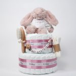 Tarta de pañales de dos pisos compuesta por 44 unidades marca Dodot Sensitive T2, peluche principal conejito personalizado, conjunto cepillo y peine bebé de madera personalizado con el nombre del bebé, sujeta chupetes de silicona personalizado con el nombre del bebé y juego calcetines bebé. Dimensiones altura 45cm x 26cm ancho. Color Rosa palo