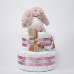 Tarta de pañales de dos pisos compuesta por 44 unidades marca Dodot Sensitive T2, peluche principal conejito personalizado, conjunto cepillo y peine bebé de madera personalizado con el nombre del bebé, sujeta chupetes de silicona personalizado con el nombre del bebé y juego calcetines bebé. Dimensiones altura 45cm x 26cm ancho. Color Rosa palo