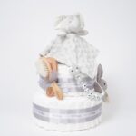 Tarta de pañales de dos pisos compuesta por 44 unidades marca Dodot, peluche principal Doudou personalizable, mordedor orejitas de crochet y madera personalizado, sujeta chupetes de silicona personalizado, conjunto de cepillo y peine bebé personalizados. Dimensiones altura 40cm x 26cm ancho. Color Gris