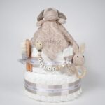 Tarta de pañales de dos pisos compuesta por 44 unidades marca Dodot, peluche principal Doudou personalizable, mordedor orejitas de crochet y madera, sujeta chupetes de silicona personalizado, conjunto de cepillo y peine bebé personalizados. Dimensiones altura 40cm x 26cm ancho. Color Toffe