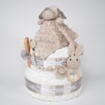 Tarta de pañales de dos pisos compuesta por 44 unidades marca Dodot, peluche principal Doudou personalizable, mordedor orejitas de crochet y madera, sujeta chupetes de silicona personalizado, conjunto de cepillo y peine bebé personalizados. Dimensiones altura 40cm x 26cm ancho. Color Toffe