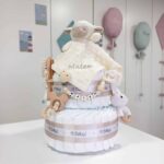 Tarta de pañales de dos pisos compuesta por 44 unidades marca Dodot Sensitive T2, peluche principal osito Dou Dou personalizado, mordedor orejitas de crochet y madera, sujeta chupetes de silicona personalizado, conjunto de cepillo y peine bebé de madera personalizados. Dimensiones altura 40cm x 26cm ancho. Color Beige