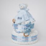Tarta de pañales de dos pisos compuesta por 44 unidades marca Dodot Sensitive T2, peluche principal osito Dou Dou personalizado, mordedor orejitas de crochet y madera, sujeta chupetes de silicona personalizado, conjunto de cepillo y peine bebé de madera personalizados. Dimensiones altura 40cm x 26cm ancho. Color Azul