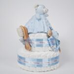 Tarta de pañales de dos pisos compuesta por 44 unidades marca Dodot Sensitive T2, peluche principal osito Dou Dou personalizado, mordedor orejitas de crochet y madera, sujeta chupetes de silicona personalizado, conjunto de cepillo y peine bebé de madera personalizados. Dimensiones altura 40cm x 26cm ancho. Color Azul