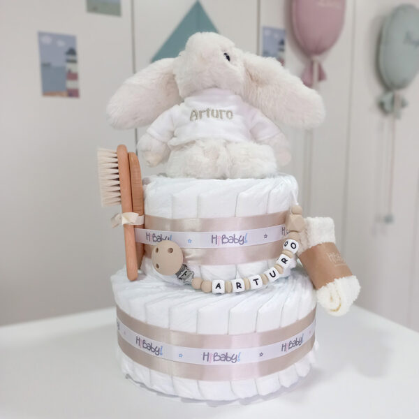 Tarta de pañales de dos pisos compuesta por 44 unidades marca Dodot Sensitive T2, peluche principal conejito personalizado, conjunto cepillo y peine bebé de madera personalizado, sujeta chupetes de silicona personalizado y juego calcetines bebé. Dimensiones altura 45cm x 26cm ancho. Color Beige/Toffe