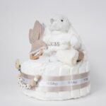 Tarta de pañales de un piso compuesto por 20 unidades marca Dodot Sensitive T2, peluche principal conejito personalizado, mordedor de piqué waffle de algodón y aro de madera personalizado, sujeta chupetes de silicona modelo Momma personalizado y juego calcetines bebé de algodón. Dimensiones altura 35cm x 22cm ancho. Color Toffe