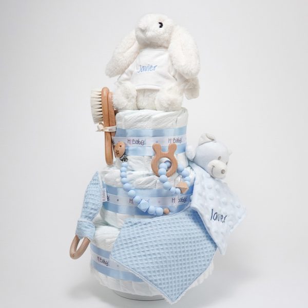 Tarta de pañales de tres pisos compuesto por 52 unidades marca Dodot sensitive, peluche principal conejito personalizado, sujeta chupetes de silicona Momma, mordedor orejitas piqué waffle algodón con aro de madera personalizado con el nombre del bebé, cepillo y peine para bebé de madera 100% natural personalizado con el nombre del bebé, mordedor de silicona Momma, bandana de piqué waffle y Doudou animalito personalizado con el nombre del bebé. Dimensiones altura 55cm x 26cm ancho. Color azul celeste