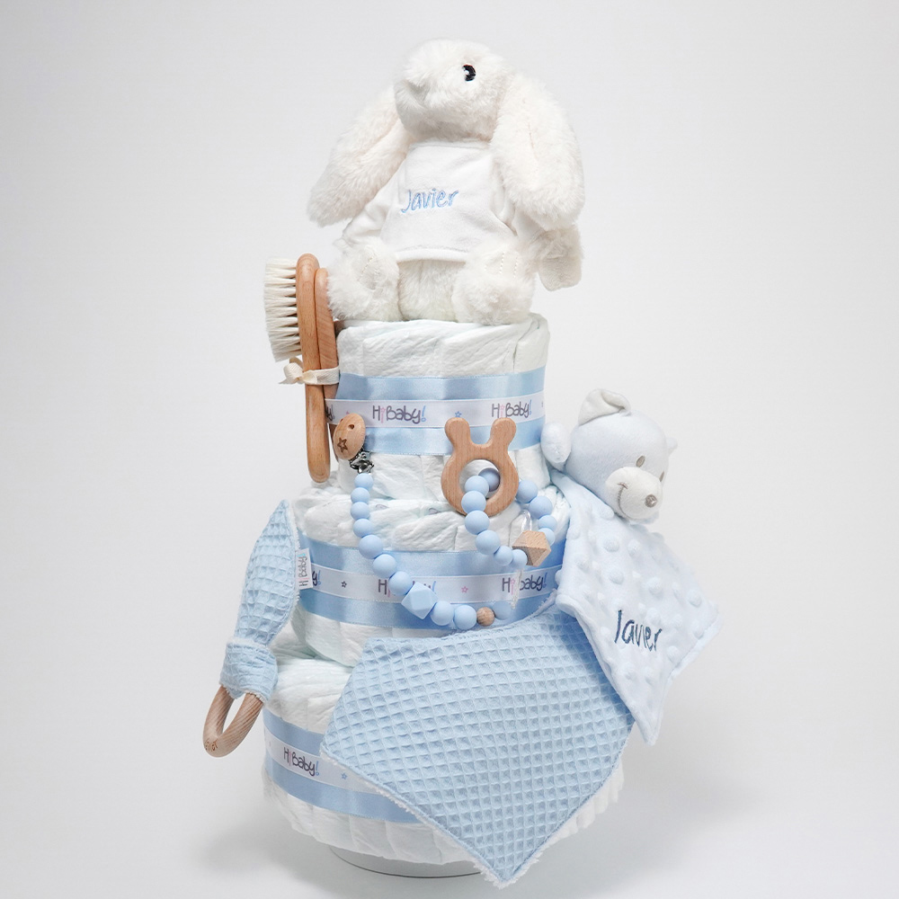 Tarta de pañales de tres pisos compuesto por 52 unidades marca Dodot sensitive, peluche principal conejito personalizado, sujeta chupetes de silicona Momma, mordedor orejitas piqué waffle algodón con aro de madera personalizado con el nombre del bebé, cepillo y peine para bebé de madera 100% natural personalizado con el nombre del bebé, mordedor de silicona Momma, bandana de piqué waffle y Doudou animalito personalizado con el nombre del bebé. Dimensiones altura 55cm x 26cm ancho. Color azul celeste