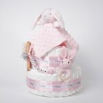 Tarta de pañales bebé de dos pisos compuesta por 44 unidades marca Dodot, peluche principal conejito Dou Dou personalizado, mordedor orejitas de crochet y madera, sujeta chupetes de silicona personalizado, conjunto de cepillo y peine bebé de madera personalizados. Dimensiones altura 40cm x 26cm ancho. Color rosa bebé