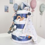 Tarta de pañales de 3 pisos compuesto por 52 unidades marca Dodot sensitive, pelota sensorial Montessori de Little Dutch, chupetero de silicona con pinza de silicona, mordedor de silicona con figura de madera natural de haya, muñeco JIMMY de Little Dutch 35cm., Chupetero de tela Sailors bay de Little Dutch, Doudou Sailors bay de Little Dutch, stick de madera personalizado con el mnombre del bebé. Dimensiones altura 42cm x 27cm ancho. Color Azul jean
